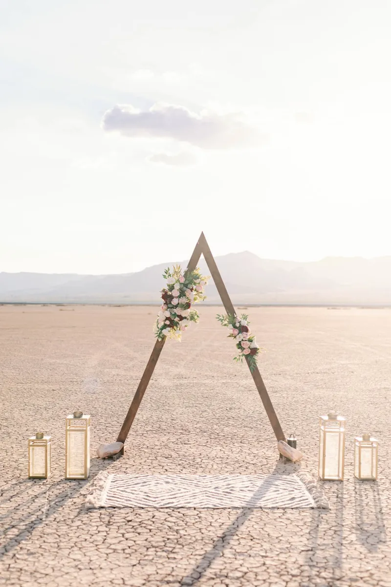 Wooden triangle arch with flowers, gold lanterns and a woven rug set up alone on the cracked lake bed