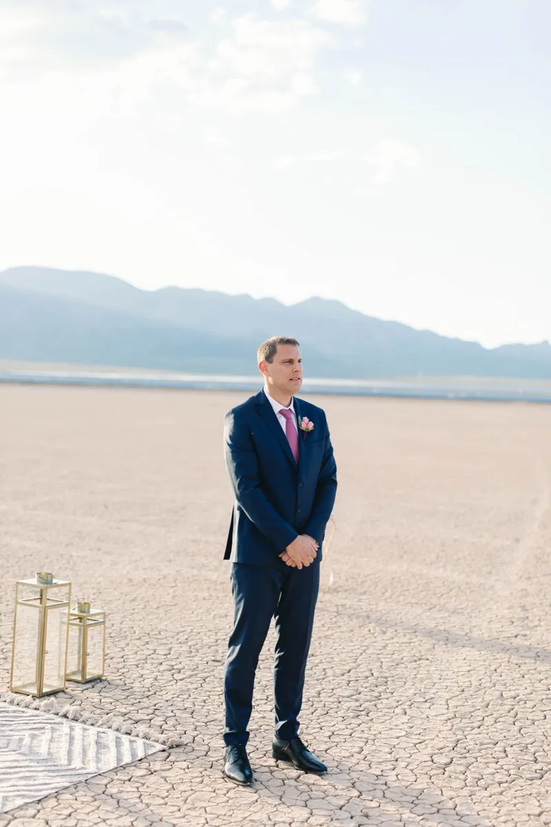 Groom in a navy suit standing alone on the lake bed before the ceremony, mountains low behind him