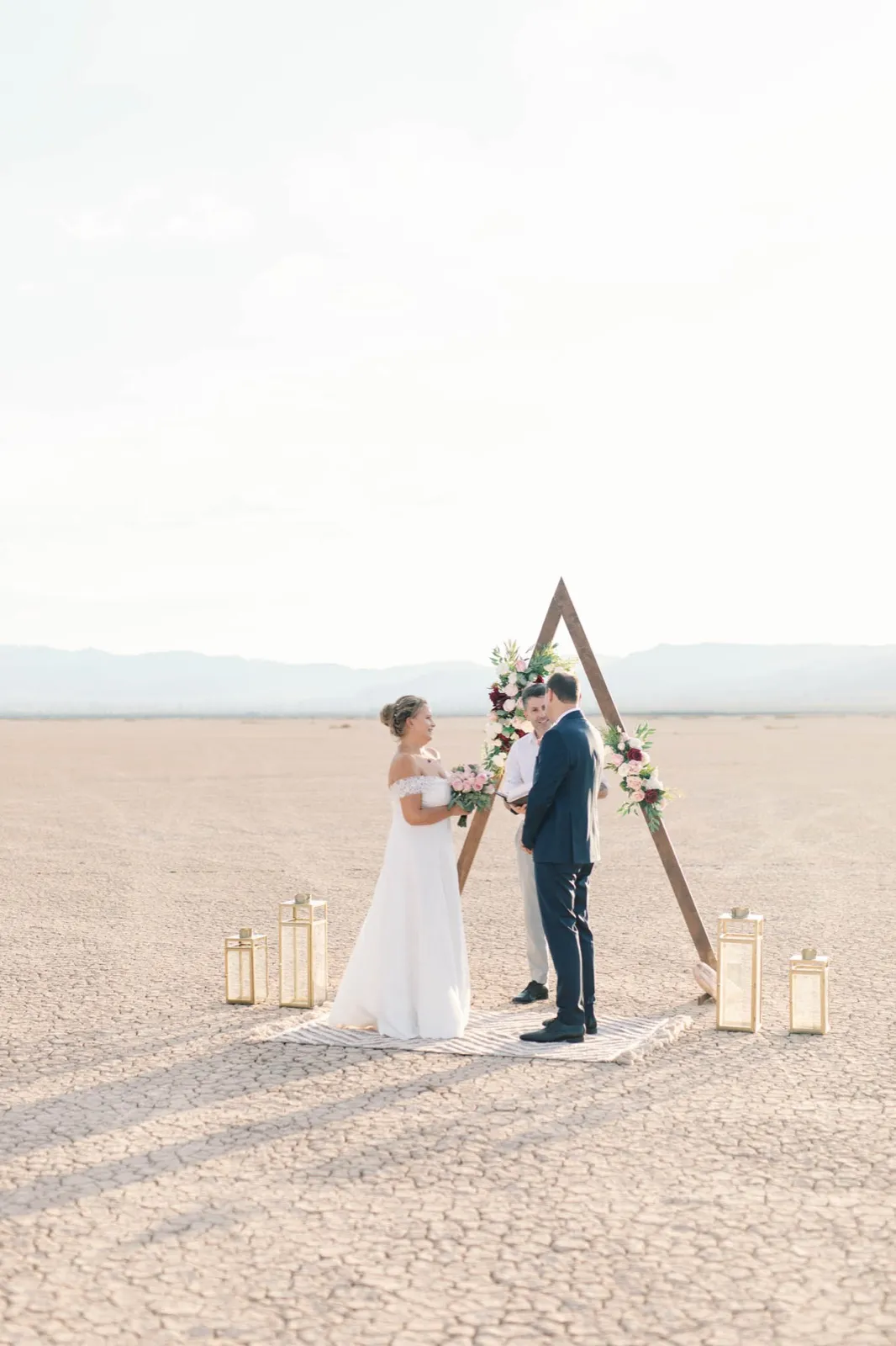 Bride and groom standing still facing each other under a flower-topped triangle arch on a cracked dry lake bed