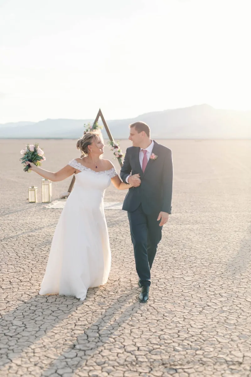 Bride holding her bouquet up and laughing as she walks arm in arm with her groom across the lake bed