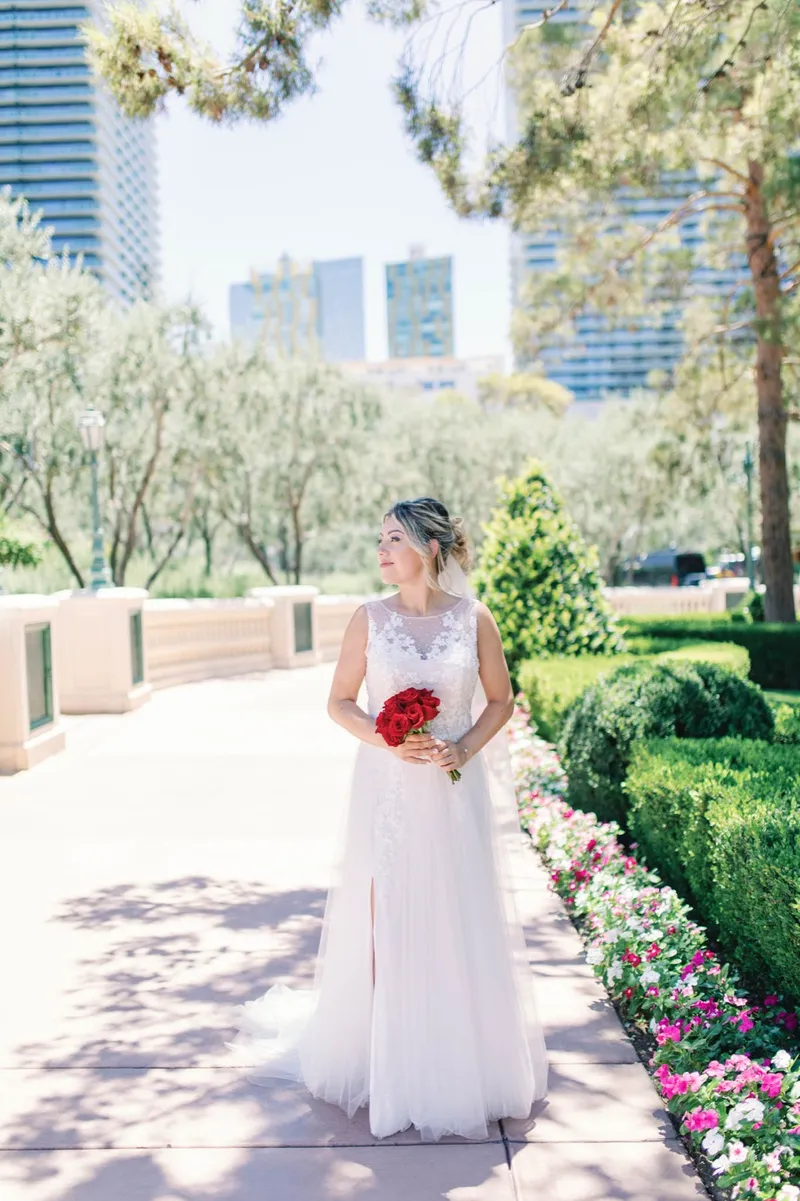 Bride in a white lace gown holding a red rose bouquet on the flower lined walkway
