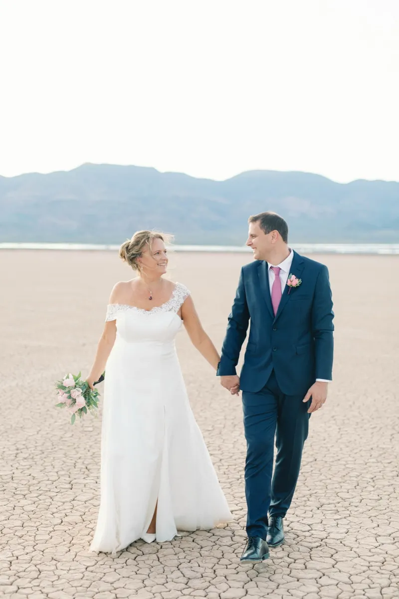 Bride and groom holding hands and walking together across the pale flat with mountains in the distance