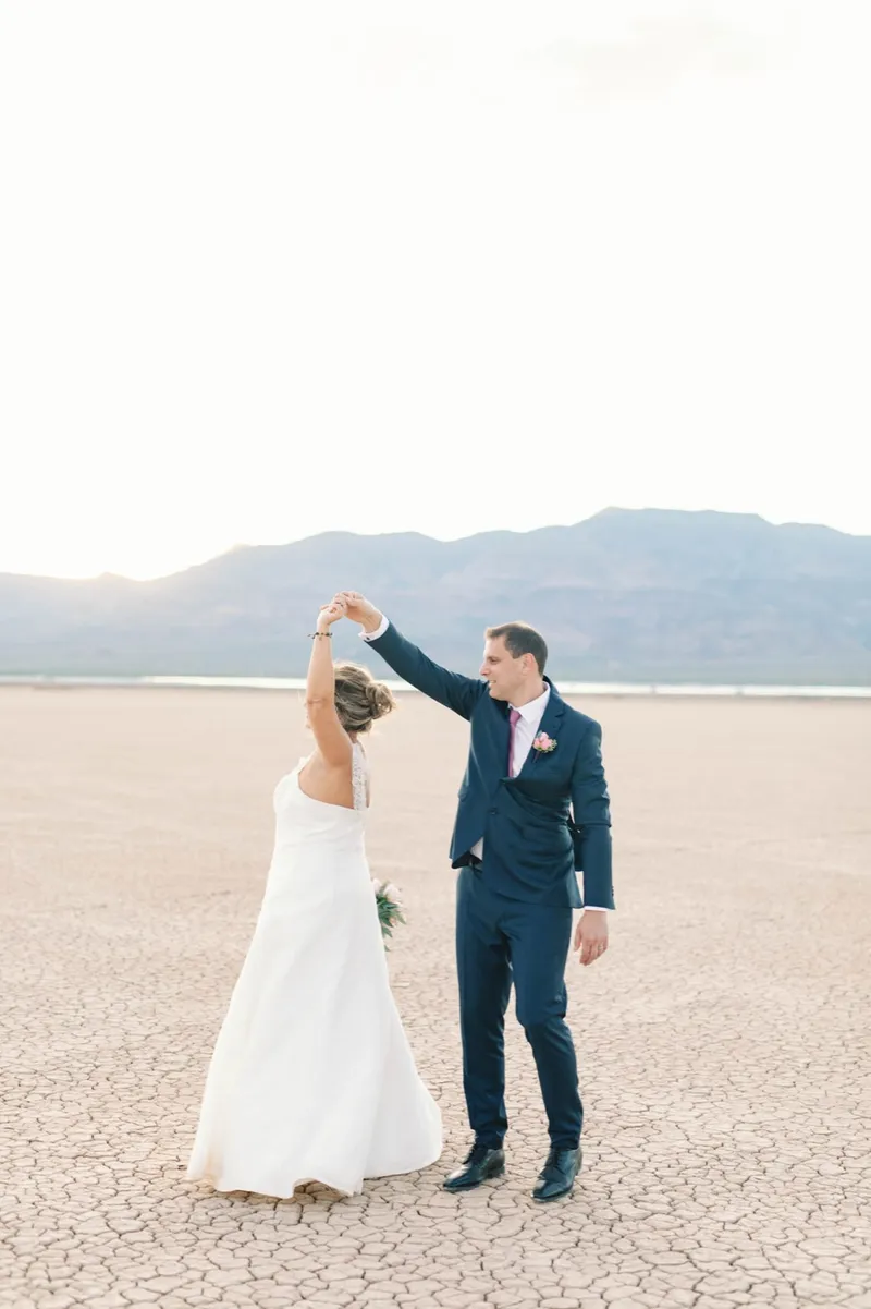 Groom in a navy suit spinning his bride under his arm on the open lake bed at sunset