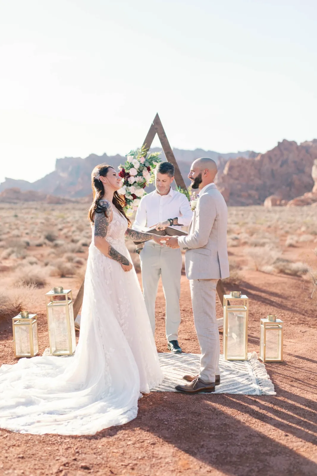 Officiant reading from an open book as a bride and groom hold hands at a triangle arch on red desert dirt
