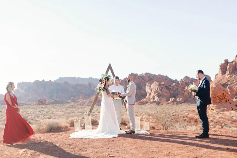 Bride and groom at a wooden triangle arch with gold lanterns while their officiant reads, two guests watching in the desert