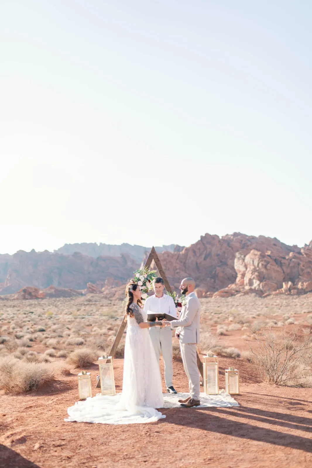 Bride and groom holding hands at a flower topped triangle arch while an officiant reads, red rock ridge behind