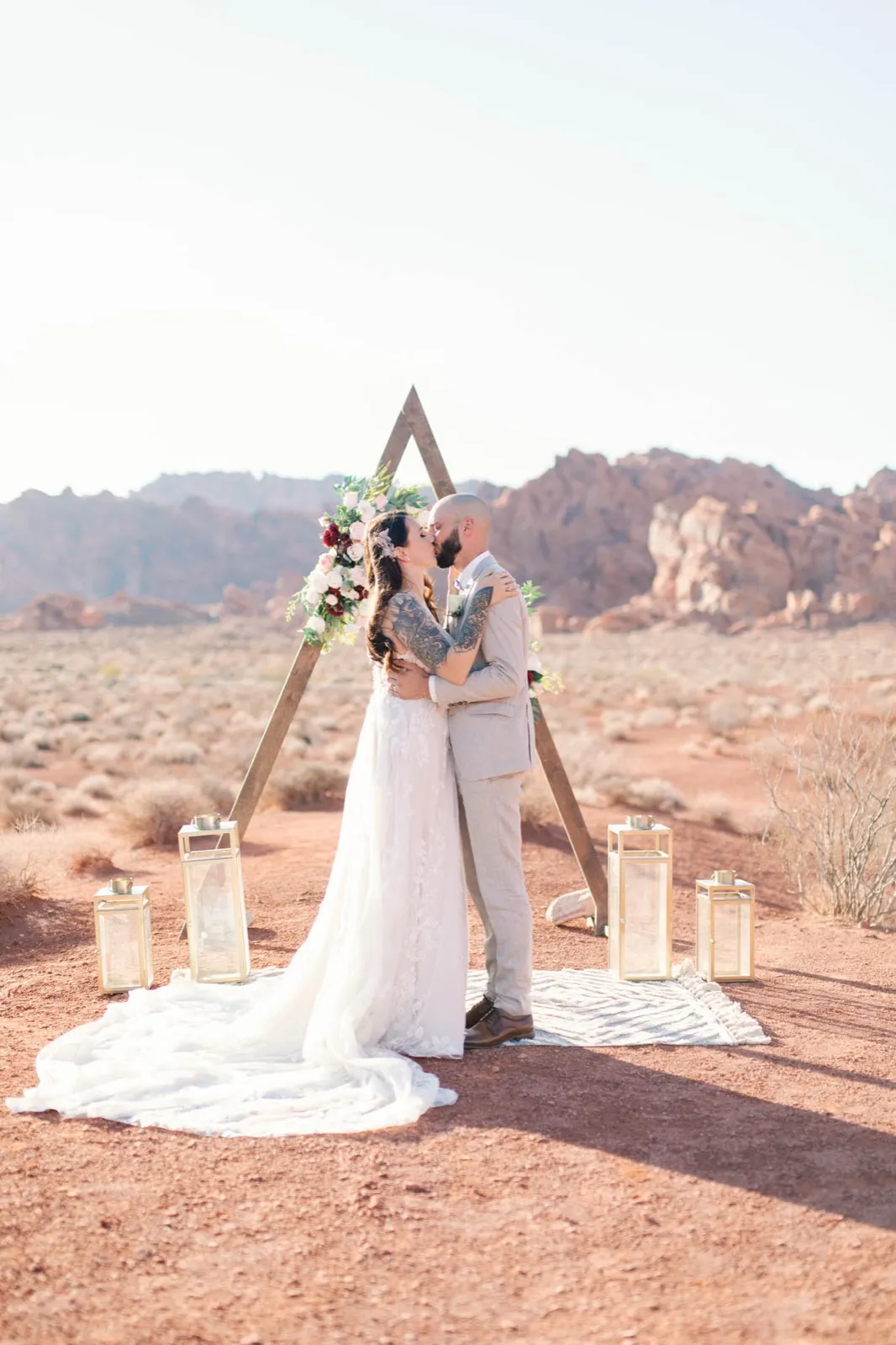 Couple kissing under a wooden triangle arch with five gold lanterns and a woven rug set on bare red dirt