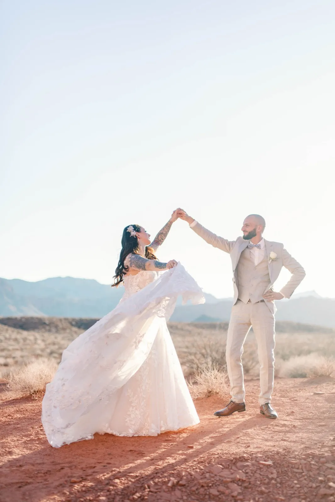 Groom spinning his bride on the red desert floor in low backlit sun, her lace skirt swinging, mountains behind