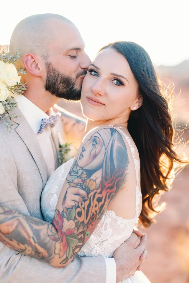 Groom kissing his brides temple as the sun lights up her tattooed arm