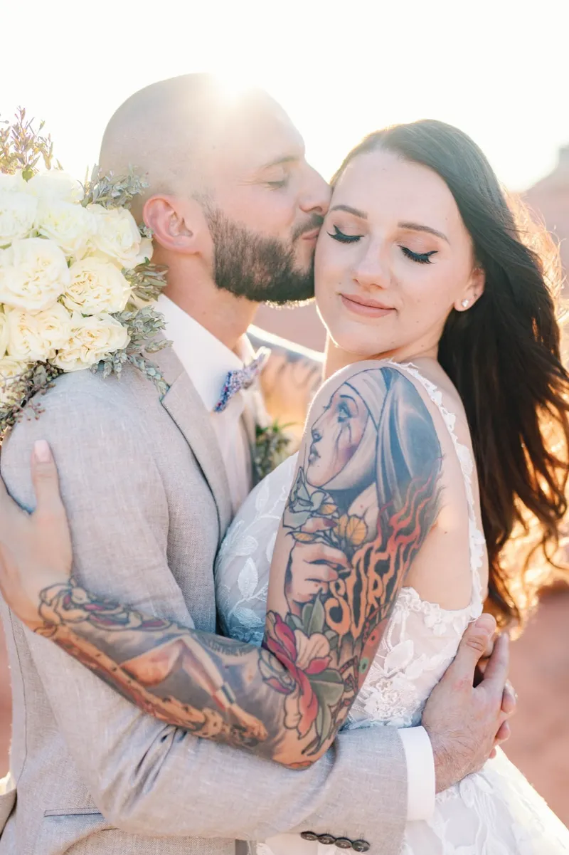 Groom kissing his bride's forehead with white roses at her shoulder