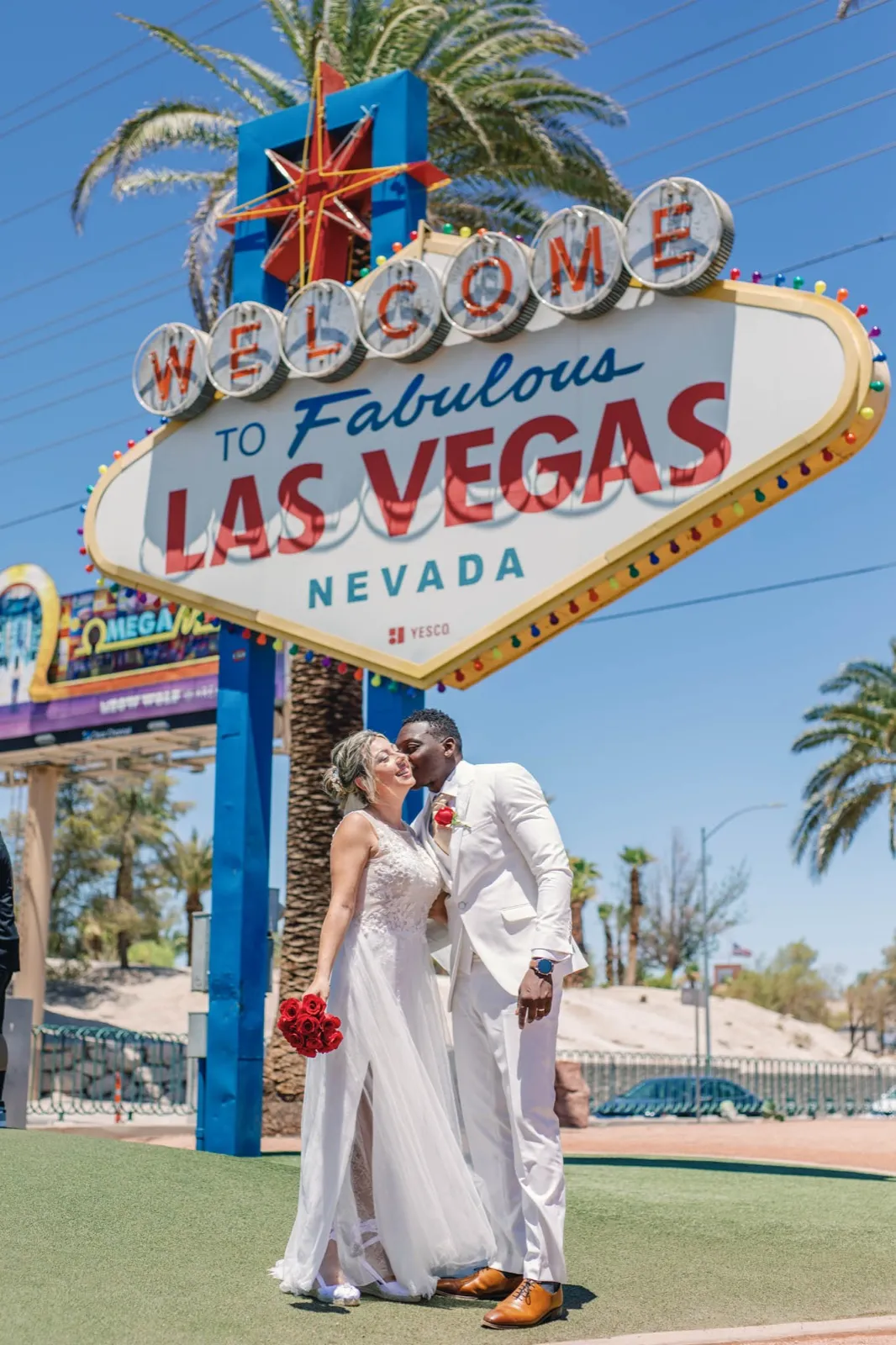 Groom in a white suit kisses his bride’s cheek on the turf under the Las Vegas sign in hard midday sun