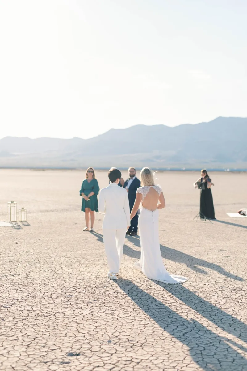 Two brides in white standing with their small group of guests out on the open lake bed