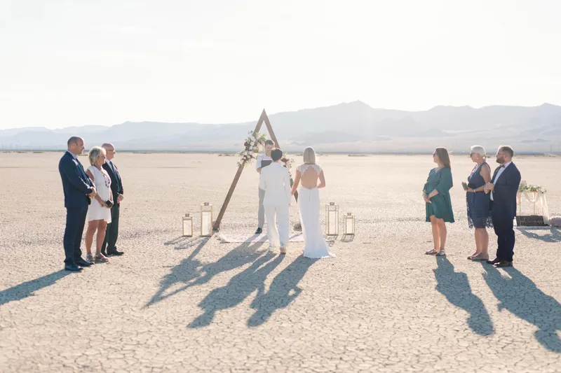 Two brides walking toward their triangle arch with long shadows stretching across the cracked ground