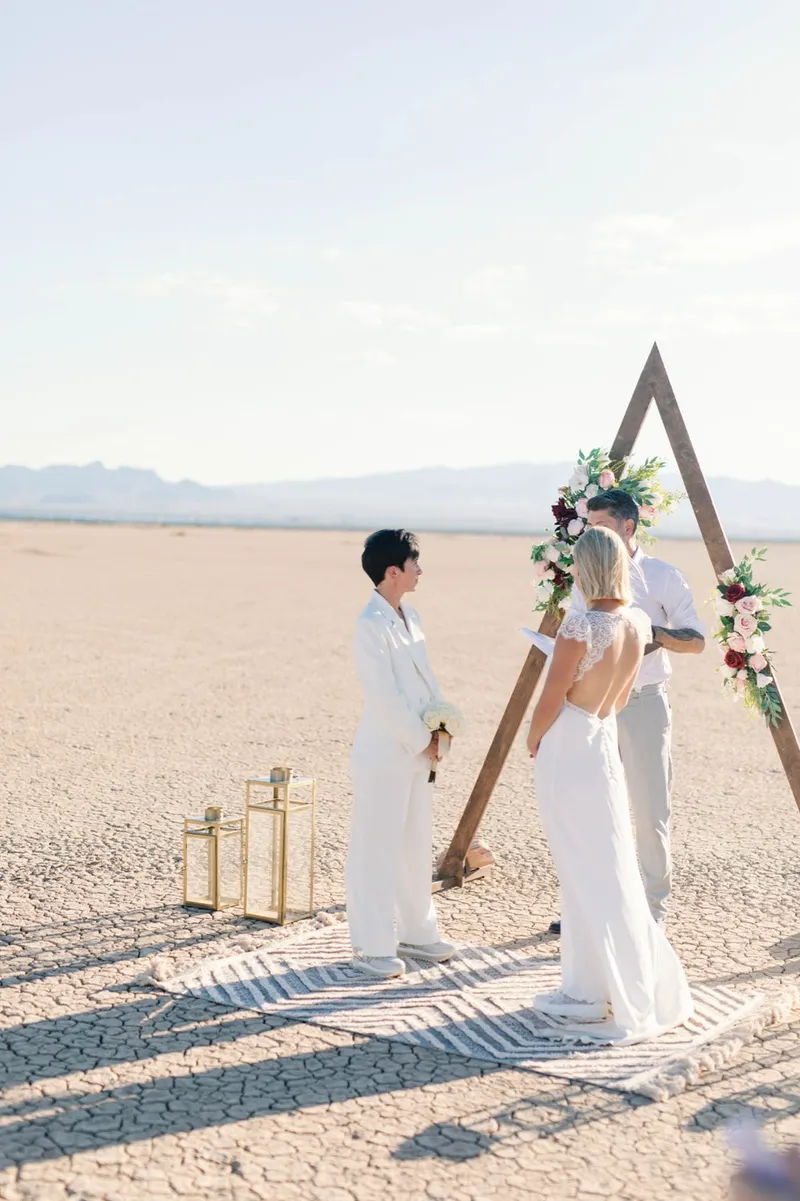 Two brides holding hands at the arch during their ceremony, one in a white suit and one in lace