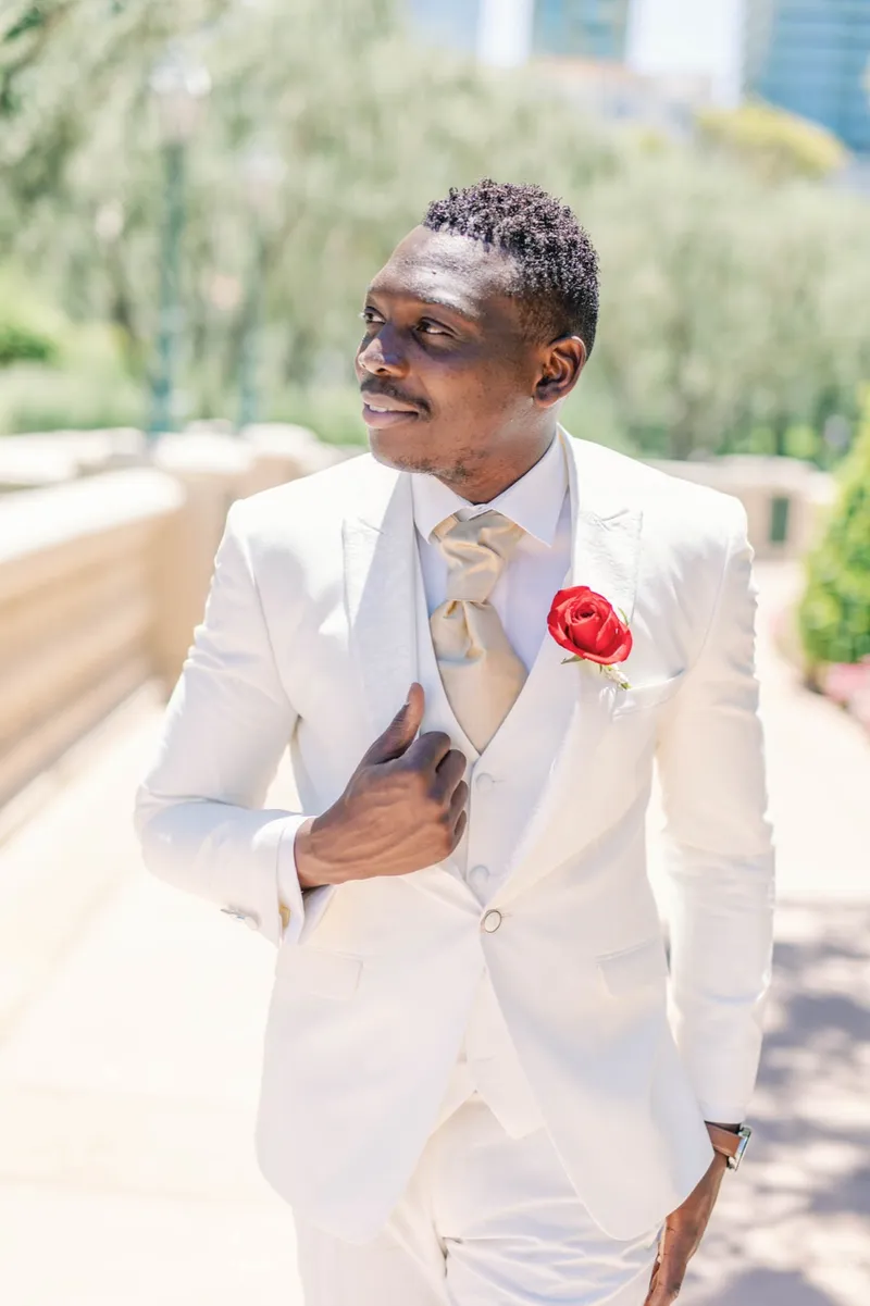 Groom in a white three piece suit with a red rose on his lapel looking off toward the fountains