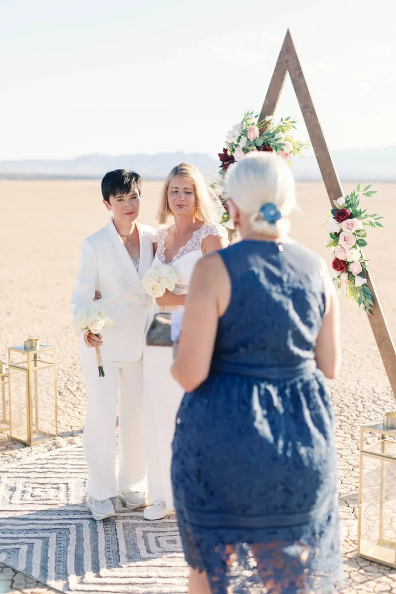 Two brides holding white bouquets as their officiant reads in front of the flower covered arch