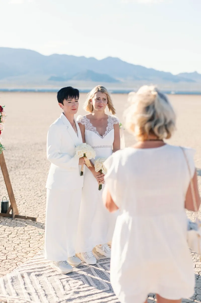 Bride in a white suit and bride in lace listening to their officiant during the ceremony