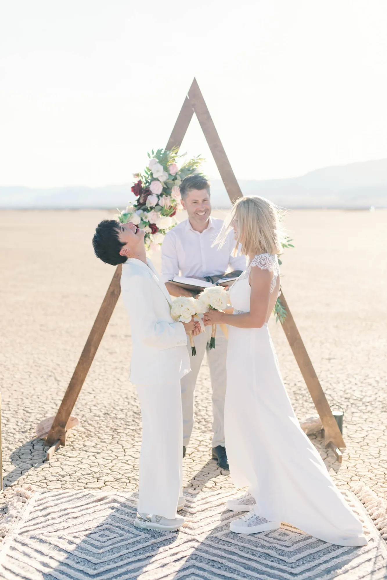 Two brides laughing with their officiant during a ceremony under a triangle wooden arch on a dry lake bed
