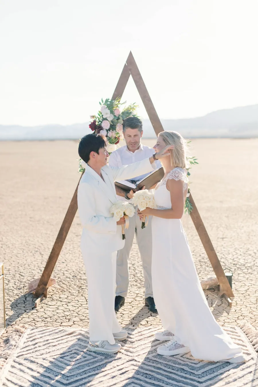 Two brides holding bouquets at their triangle arch ceremony on the dry lake bed with their officiant