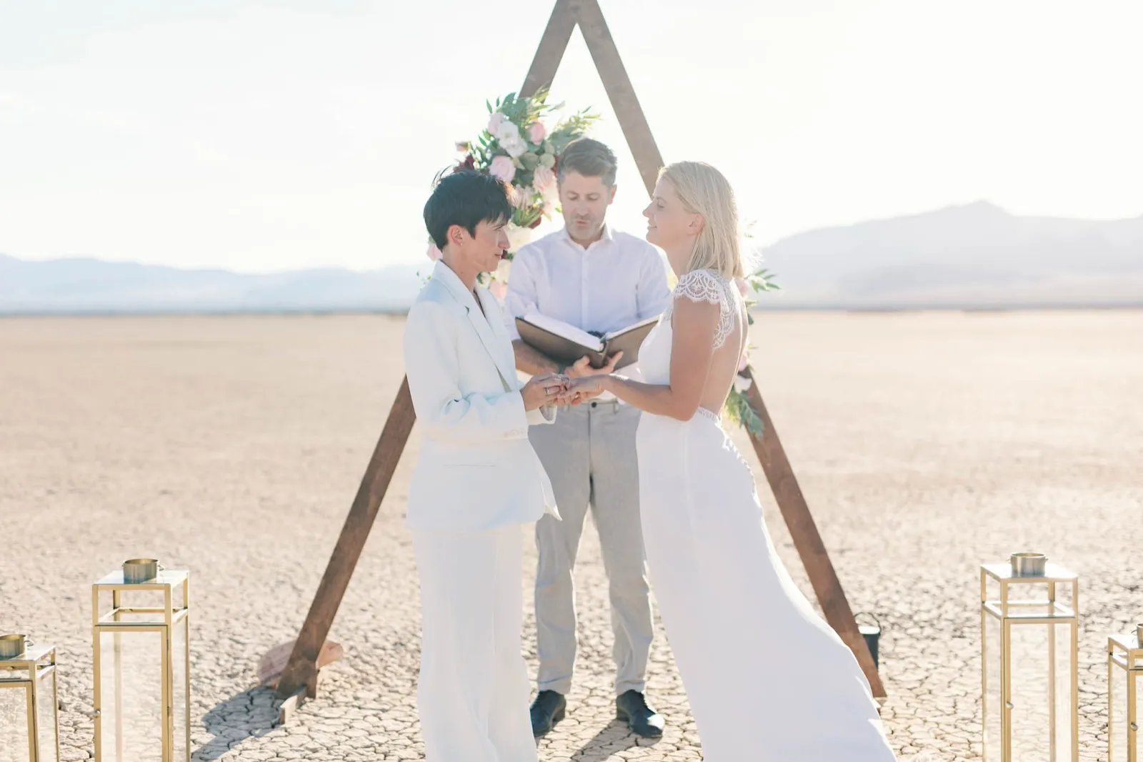 Two brides in white, one in a suit and one in a lace gown, exchanging rings as their officiant reads