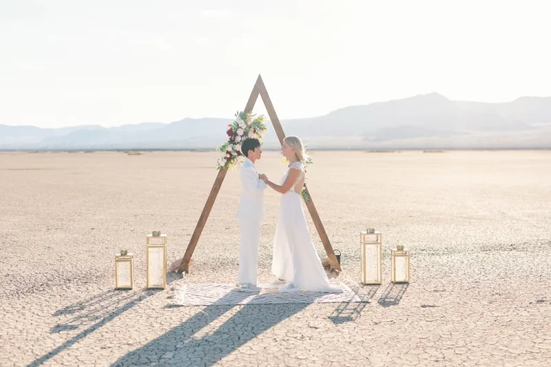 Two brides facing each other and holding both hands under the arch as they say their vows
