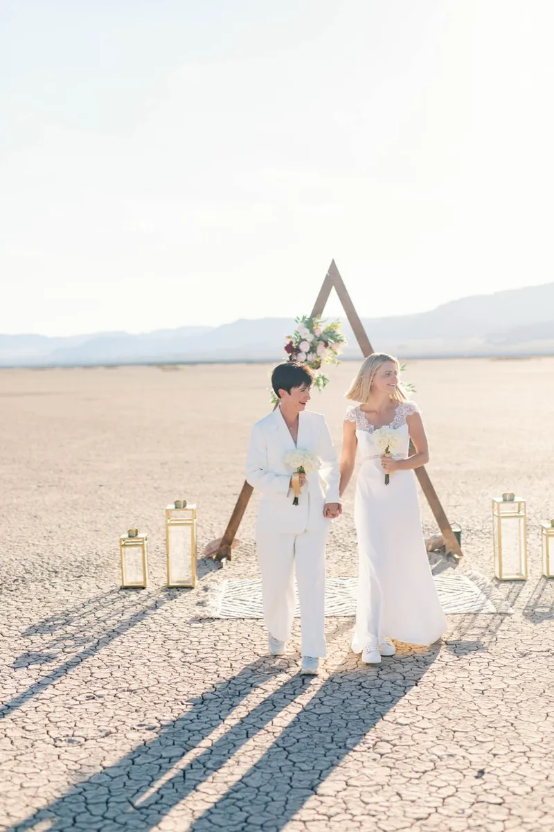 Two brides in white standing close together at the arch with the mountains far behind
