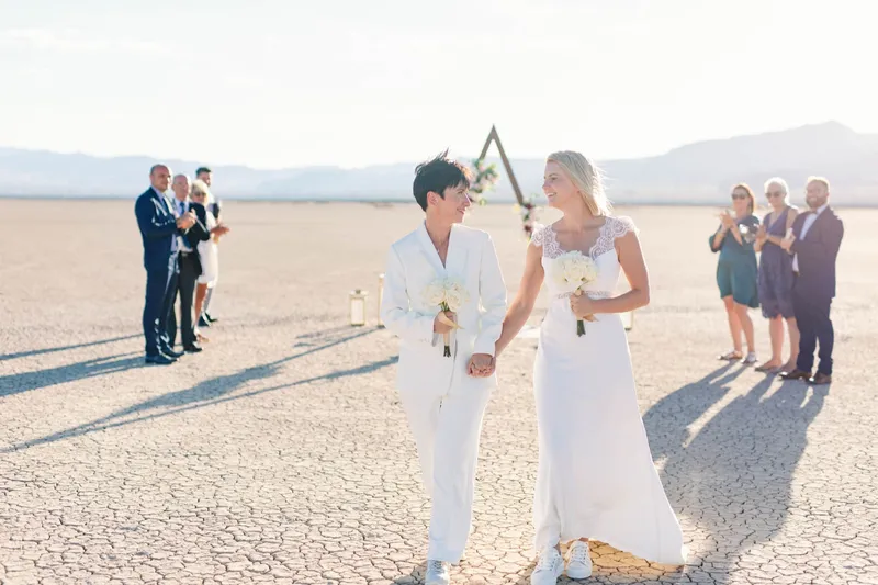 Two brides in white walking back up the aisle holding hands while their guests clap on either side