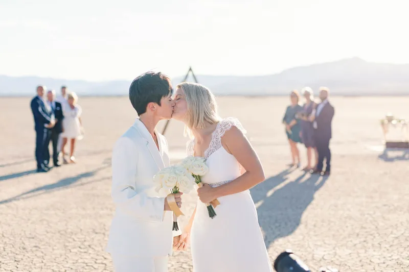 Two brides kissing after their ceremony while their guests watch from behind