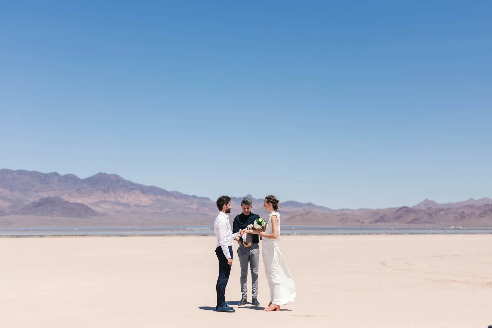 Couple standing with their officiant alone in the middle of a pale cracked lake bed with a long mountain range behind them