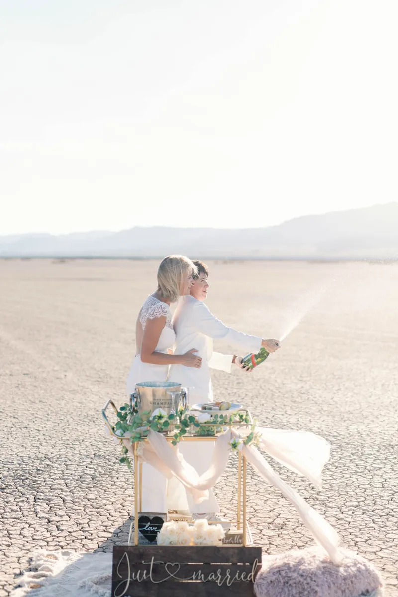 Two brides spraying a bottle of champagne beside a gold cart and just married sign on the dry lake bed