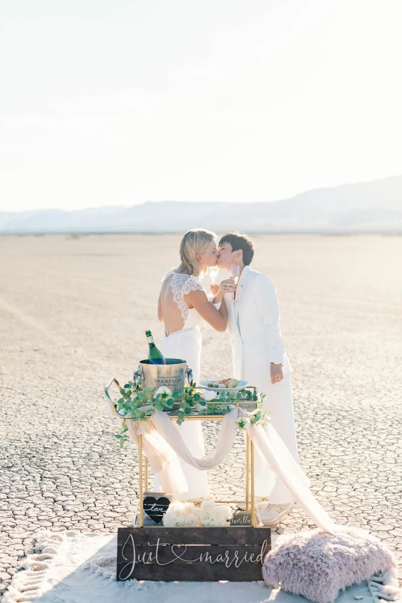 Two brides sharing a drink and a kiss beside their champagne cart on the flat