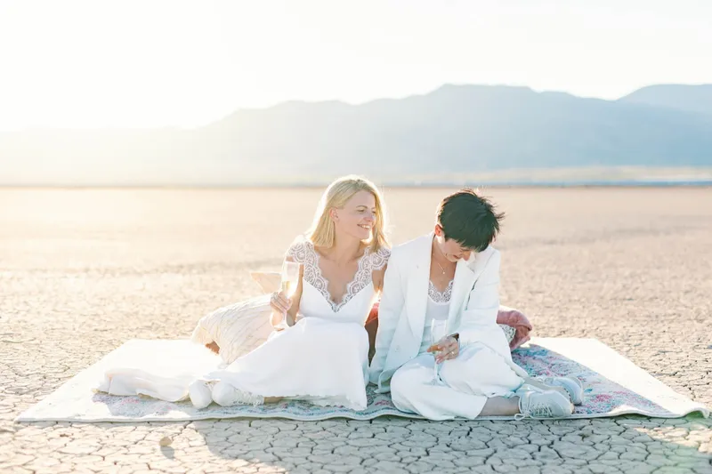 Two brides sitting together on a rug on the lake bed with champagne glasses in their hands