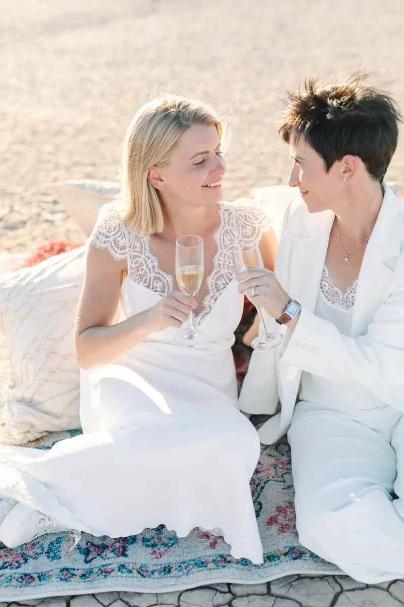 Two brides sitting on cushions with champagne, leaning into each other and smiling