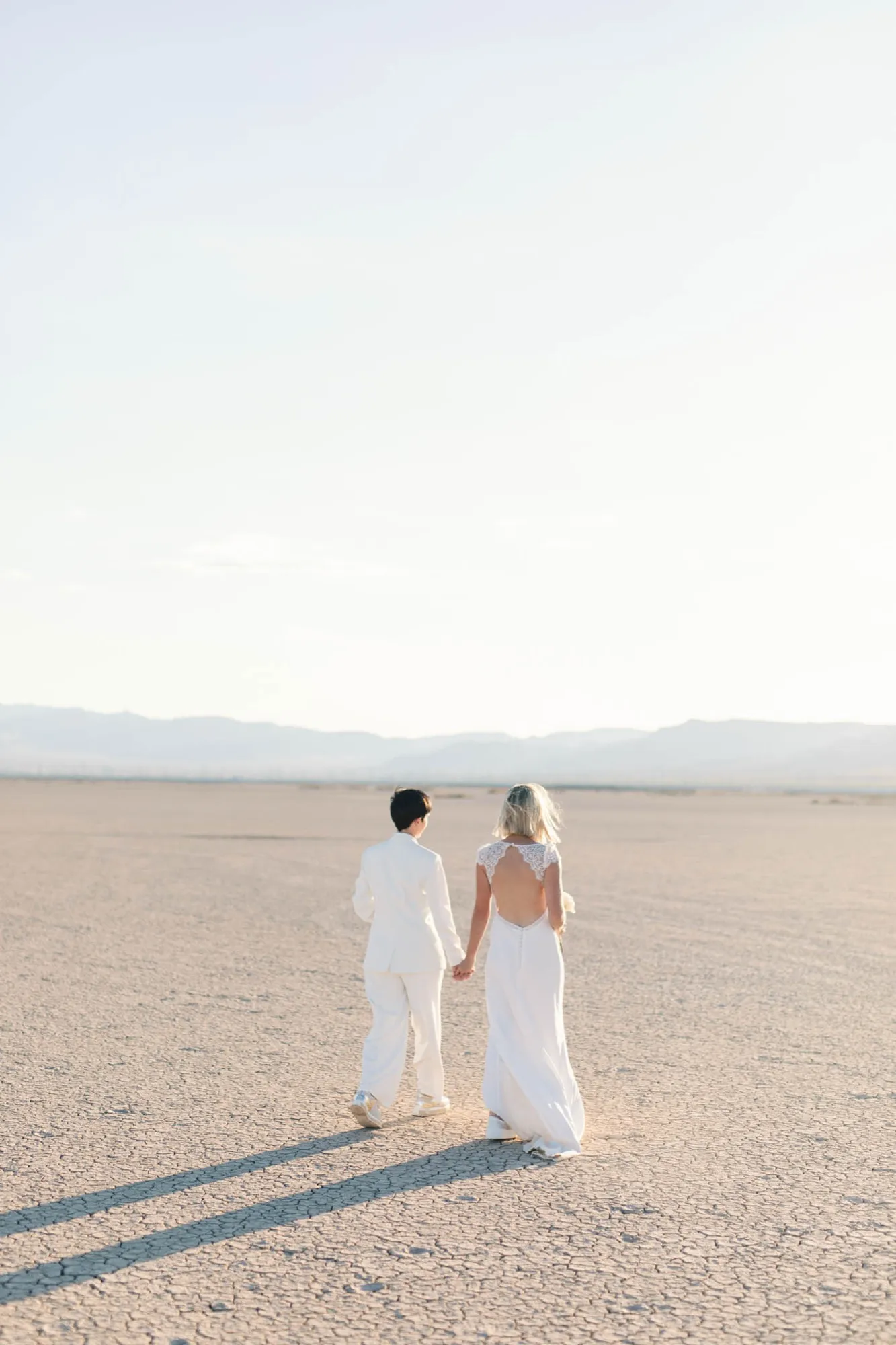 Two brides walking hand in hand across the cracked earth of a dry lake bed, mountains far in the distance