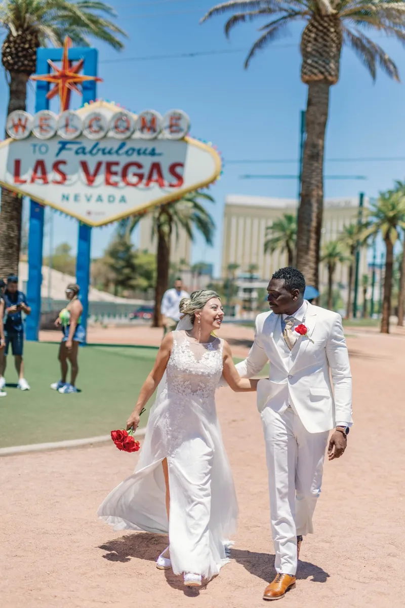A bride with red roses and her groom in a white suit walking below the Welcome to Fabulous Las Vegas sign