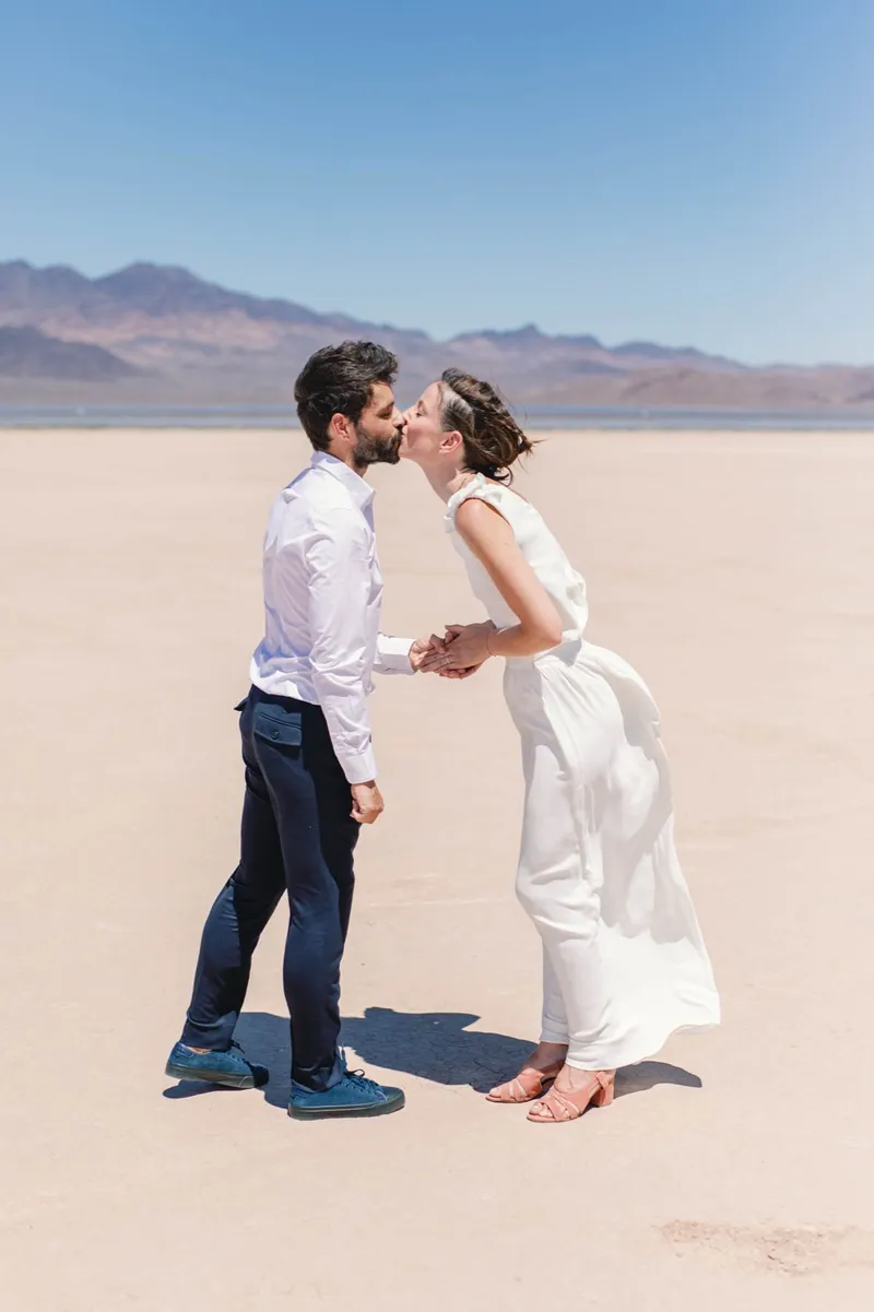 Groom in a white shirt kissing his bride as her skirt lifts in the wind on the lake bed