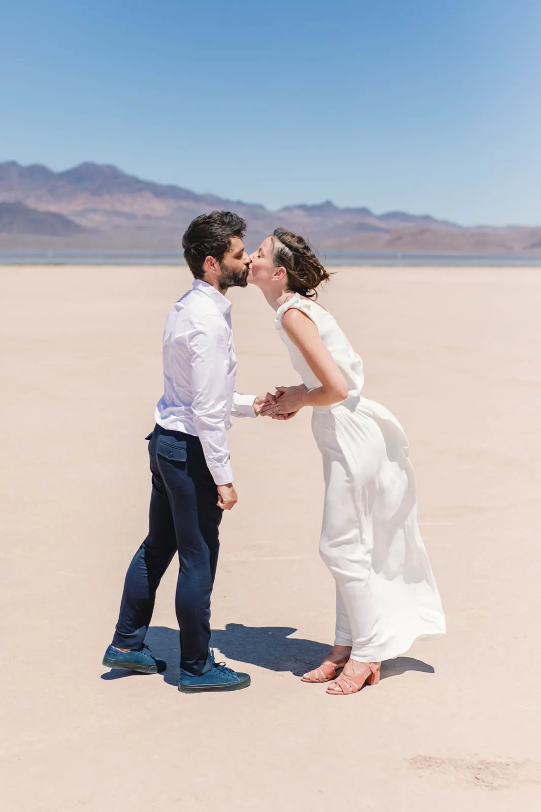 Groom in a white shirt kissing his bride as her skirt lifts in the wind on a pale cracked dry lake bed