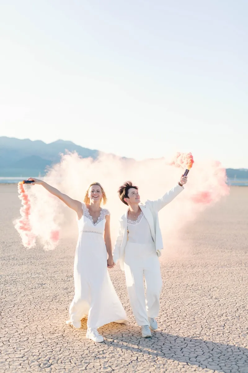 Two brides holding pink smoke bombs out wide as the smoke drifts across the flat