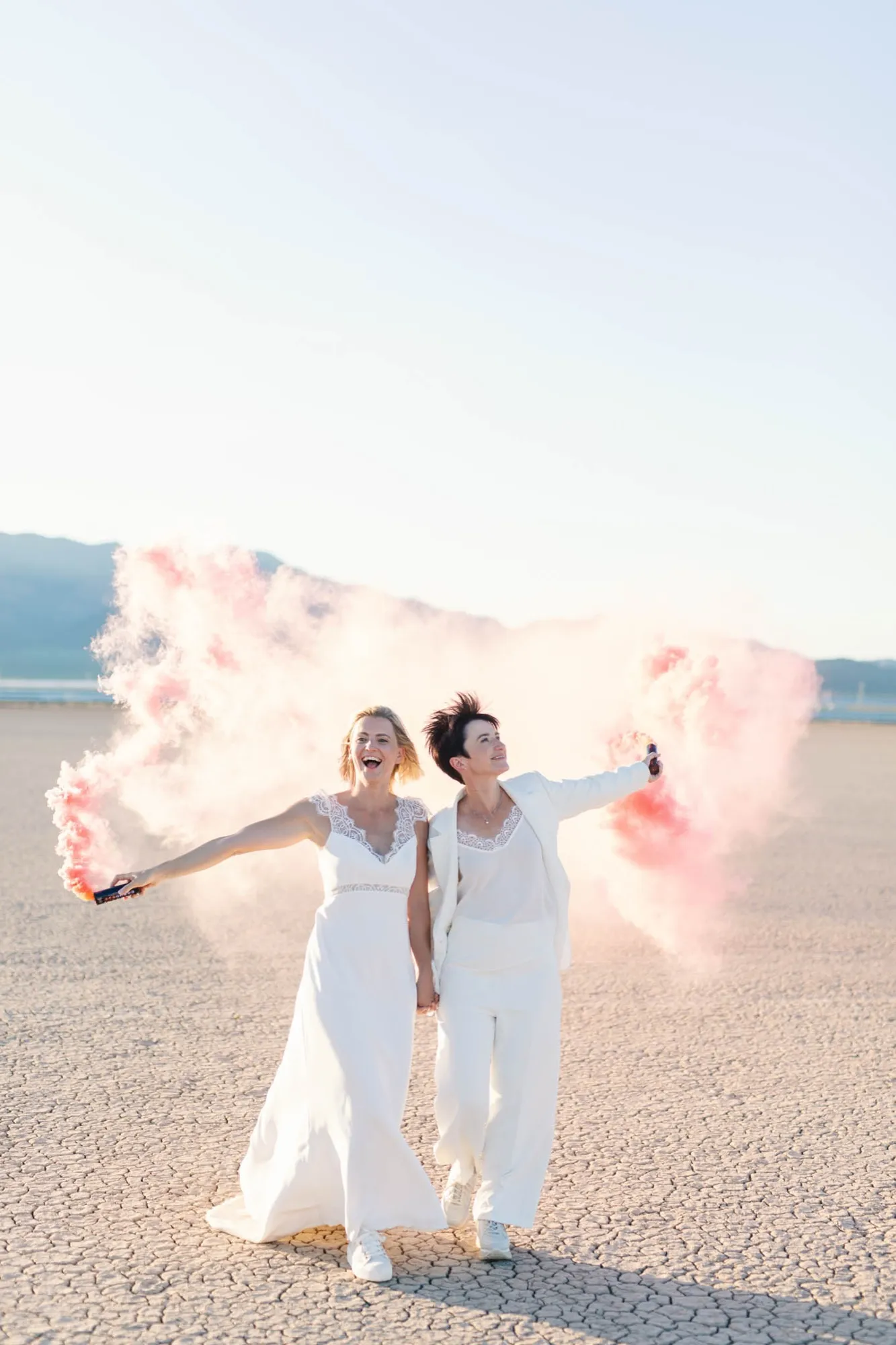 Two brides holding hands and waving pink smoke flares across the dry lake bed outside Las Vegas