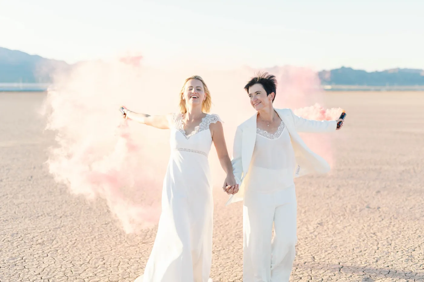 Two brides in white holding hands and waving pink smoke on the dry lake bed outside Las Vegas