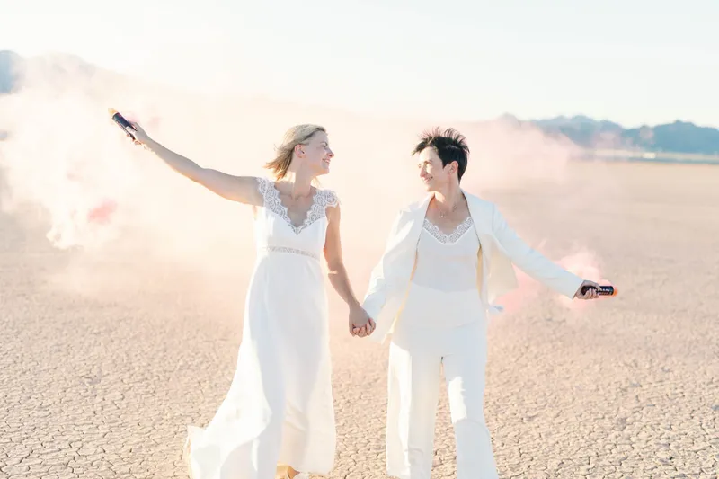 Two brides walking and laughing with pink smoke pouring from smoke bombs in their outstretched hands
