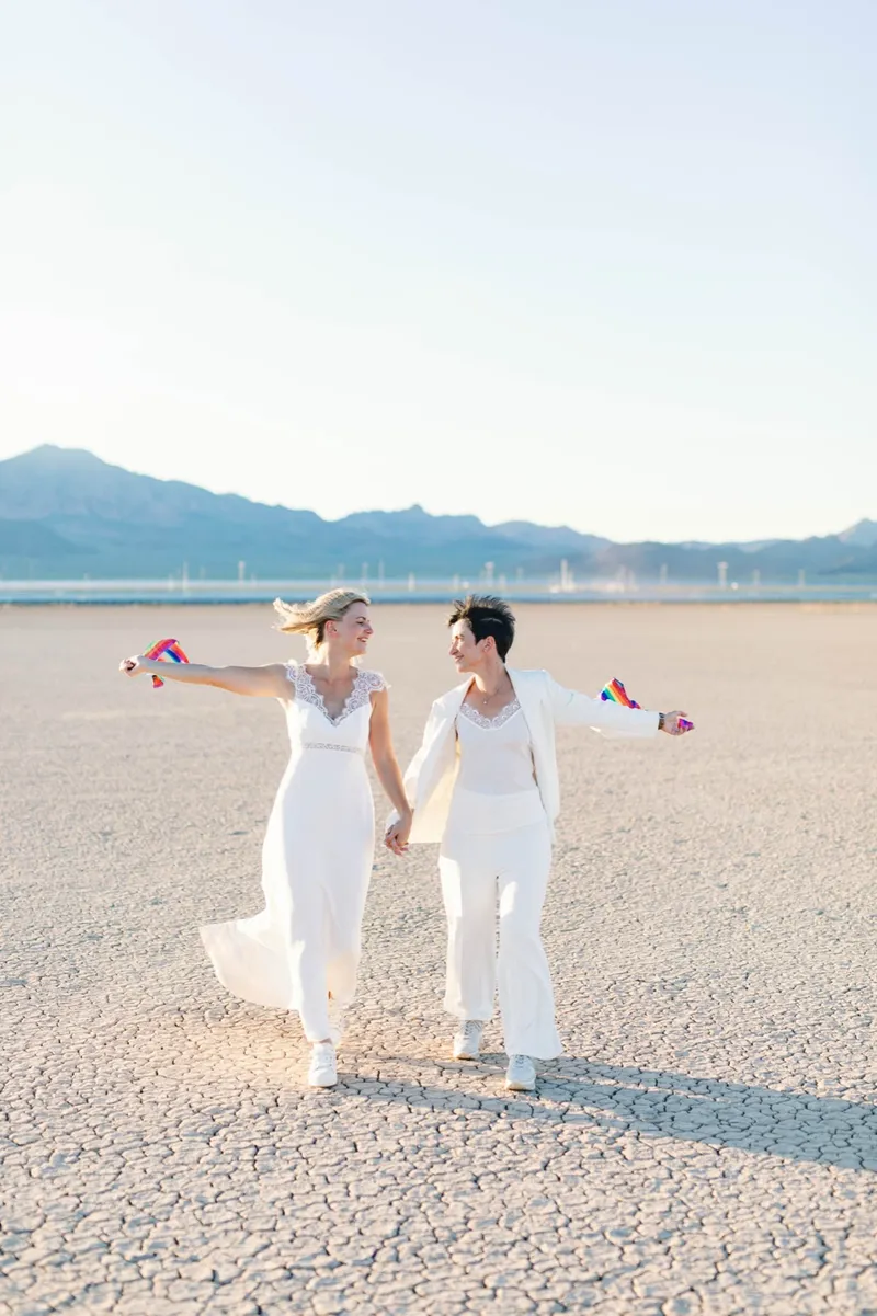 Two brides running hand in hand across the lake bed with small rainbow flags streaming from their fingers