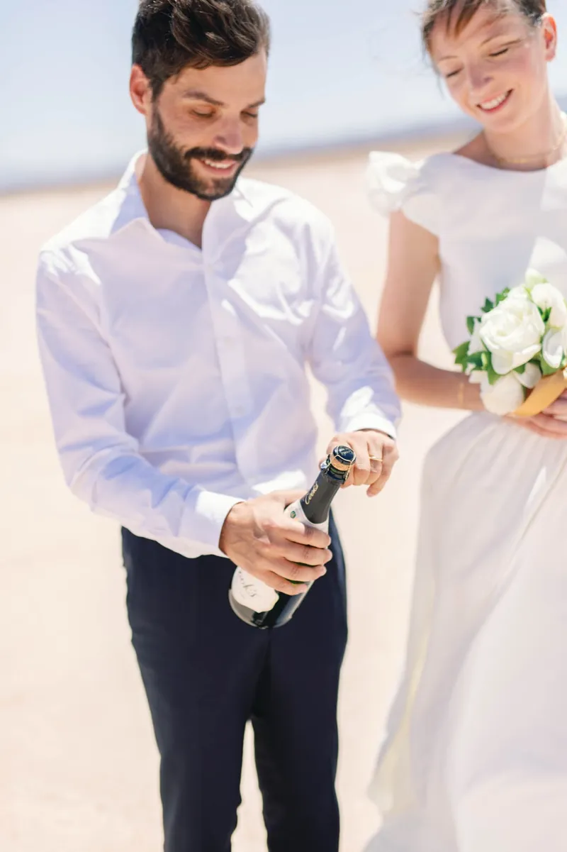 Groom opening a bottle of champagne beside his bride and her white bouquet