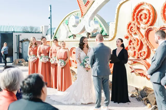 Bride and groom facing their officiant during a ceremony in front of old neon signs, bridesmaids in rust colored dresses standing alongside