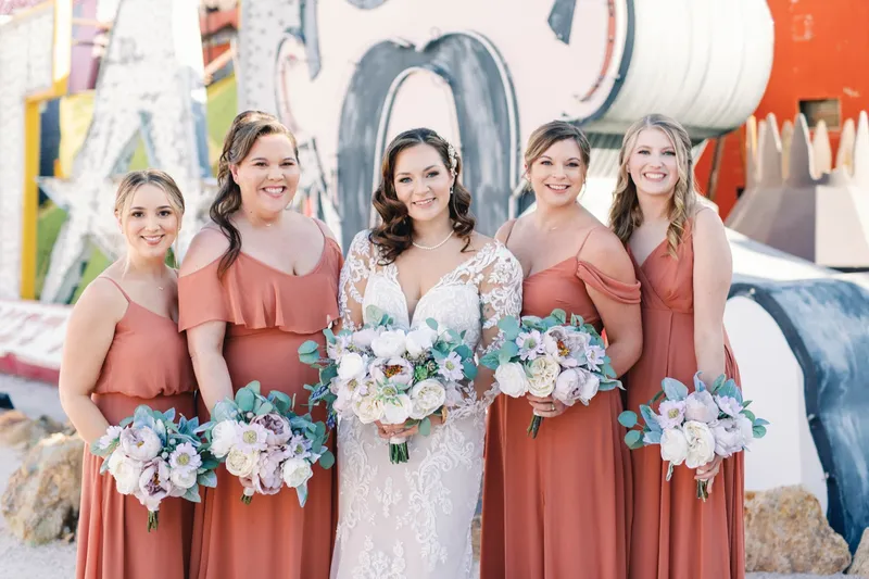 Bride and her bridesmaids in rust coloured gowns holding pale bouquets in front of the signs