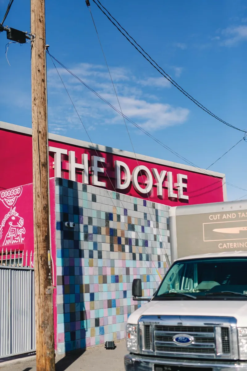 The Doyle's name in raised letters on a hot pink wall above a panel of small multicolored tiles, with a wooden power pole and blue sky above it