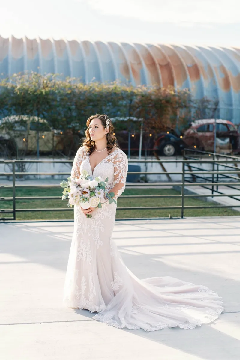 Bride standing alone on the concrete courtyard with her train spread out behind her, holding a bouquet of white flowers and eucalyptus