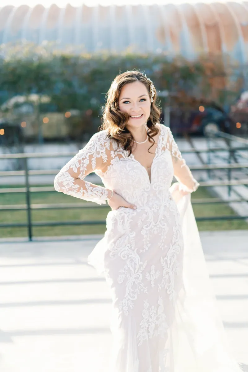 Bride laughing with a hand on her hip in her long sleeve lace gown, the hedge and the curved metal roof behind her