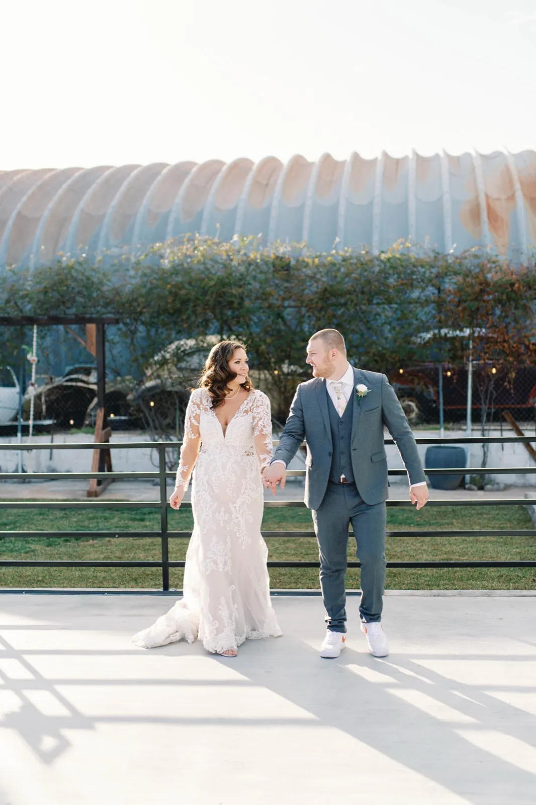 Bride and groom hold hands on a concrete courtyard in front of the long curved corrugated metal roof of a downtown venue