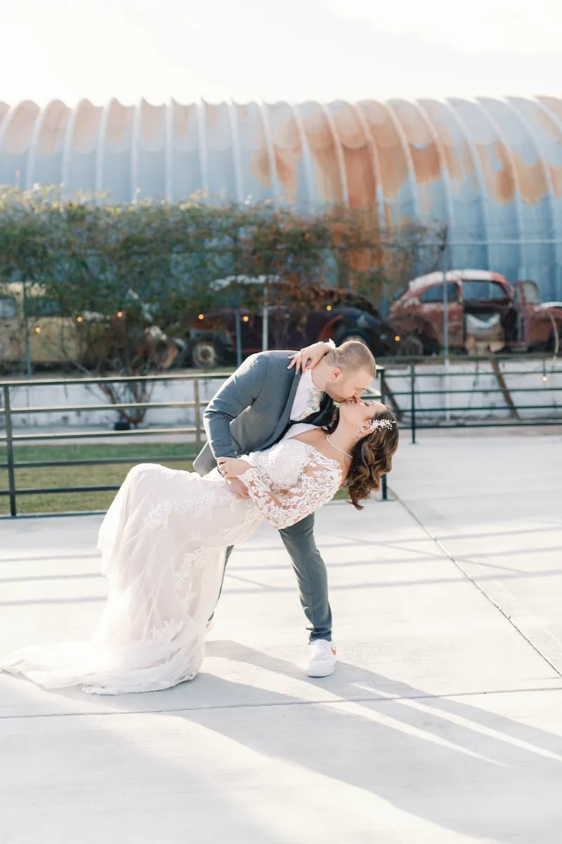Groom dipping the bride into a kiss, her train sweeping the concrete and the old cars parked along the fence behind them
